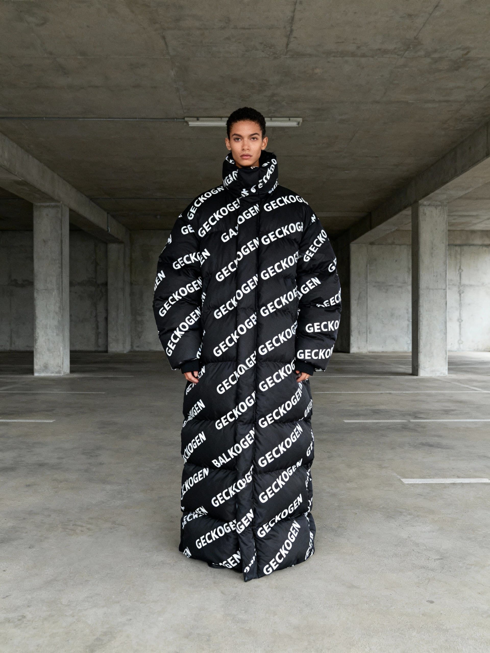 A high-fashion editorial photoshoot in Balenciaga campaign style. A striking model wears an oversized black puffer jacket completely covered in repeated "GECKOGEN" text logo pattern - the text is printed all over the jacket in various sizes creating bold graphic design, white text on black fabric. The model stands in a stark brutalist concrete environment or empty parking garage. Minimalist composition with model centered, direct confident gaze at camera. Shot on medium format camera with 50mm lens, natural desaturated color grading - muted tones, cool temperature, slightly lifted blacks. Professional fashion photography lighting - soft overcast daylight or studio setup mimicking it. The GECKOGEN text is integral to the clothing design, repeated as pattern across the entire jacket surface. Vertical portrait format. Contemporary high fashion editorial aesthetic - bold, minimal, statement-making.